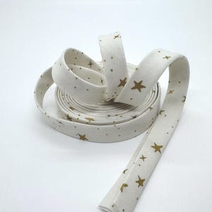 Peut inclure: Un rouleau de biais blanc cassé orné d'un motif répété d'étoiles dorées et de petits points. Le ruban est plié et prêt à être utilisé pour la couture ou les loisirs créatifs. Le ruban est en tissu.