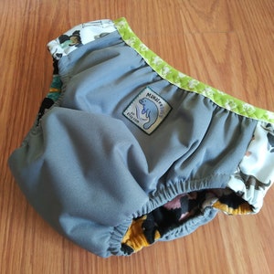 May include: Cloth diaper with a gray interior and a patterned exterior featuring cartoon animals. The diaper has a green trim with a floral pattern and a label that says "Manatee Baby".