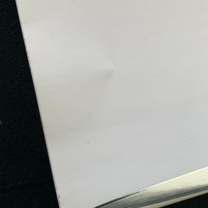 Puede incluir: Primer plano de un papel blanco con una ligera arruga y un objeto plateado en la esquina. El papel tiene una textura suave y est&aacute; sobre un fondo oscuro.