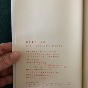 Puede incluir: Libro abierto con texto japon&eacute;s y el t&iacute;tulo "DIE T&Ouml;DLICHE DOLIS". Las p&aacute;ginas son de color crema, con texto negro y rojo. Una mano sostiene el libro abierto, revelando el texto y el dise&ntilde;o.