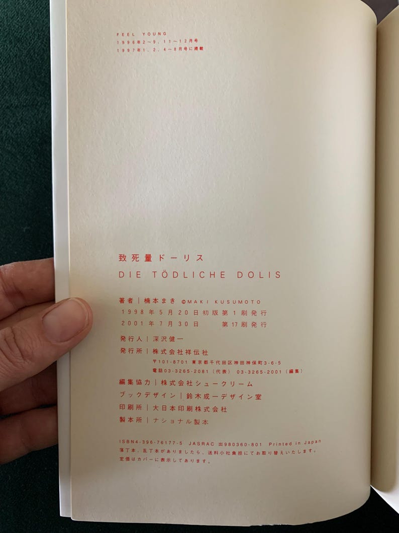 Puede incluir: Libro abierto con texto japon&eacute;s y el t&iacute;tulo "DIE T&Ouml;DLICHE DOLIS". Las p&aacute;ginas son de color crema, con texto negro y rojo. Una mano sostiene el libro abierto, revelando el texto y el dise&ntilde;o.