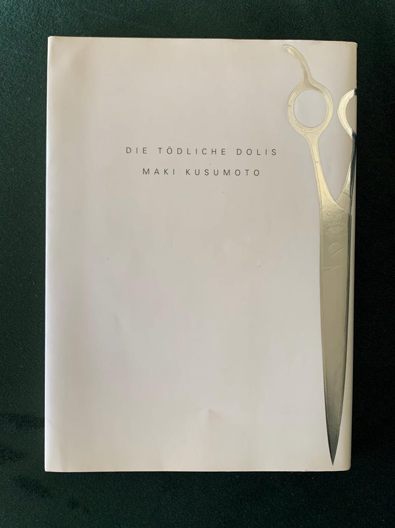 Puede incluir: Portada de libro con el t&iacute;tulo "DIE T&Ouml;DLICHE DOLIS" y el autor "MAKI KUSUMOTO" en texto negro. Un gr&aacute;fico de tijeras de peluquer&iacute;a plateadas est&aacute; en el lado derecho de la portada. El fondo es blanco.