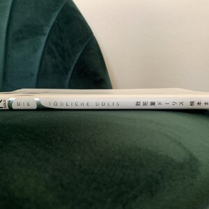 Puede incluir: Un libro blanco titulado "DIE T&Ouml;DLICHE DOLIS" con texto japon&eacute;s, apoyado en una silla de terciopelo verde oscuro. El lomo del libro es visible, mostrando el t&iacute;tulo y el nombre del autor. El libro est&aacute; en posici&oacute;n horizontal.
