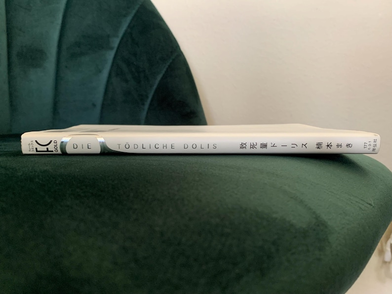 Puede incluir: Un libro blanco titulado "DIE T&Ouml;DLICHE DOLIS" con texto japon&eacute;s, apoyado en una silla de terciopelo verde oscuro. El lomo del libro es visible, mostrando el t&iacute;tulo y el nombre del autor. El libro est&aacute; en posici&oacute;n horizontal.