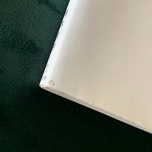 Puede incluir: Una esquina de un libro blanco con texto japon&eacute;s. El borde del libro est&aacute; ligeramente desgastado, con una peque&ntilde;a mancha marr&oacute;n. El fondo es una superficie verde oscuro, posiblemente de terciopelo.