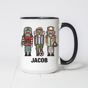 Puede incluir: Taza de cerámica blanca con asa y borde negros, con tres cascanueces de dibujos animados vestidos con uniformes deportivos. La taza tiene el nombre "JACOB" impreso en negro. Los cascanueces están vestidos como jugador de fútbol americano, fútbol y béisbol.
