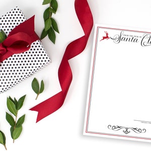 May include: A white notepad with a red border and black text that says "From the Desk of Santa Claus". There are two red reindeer illustrations on the top and bottom of the notepad. A red ribbon, a black and white polka dot gift box with a red bow, and green leaves are in the background.