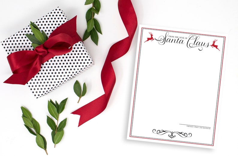 May include: A white notepad with a red border and black text that says "From the Desk of Santa Claus". There are two red reindeer illustrations on the top and bottom of the notepad. A red ribbon, a black and white polka dot gift box with a red bow, and green leaves are in the background.