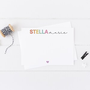 May include: A white card with the name "STELLA marie" written in rainbow colors. A small purple heart is in the bottom center of the card.