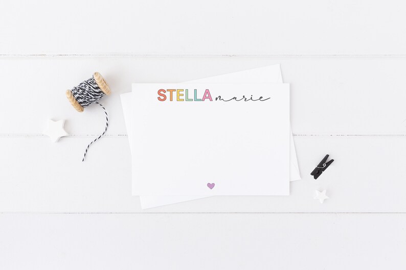 May include: A white card with the name "STELLA marie" written in rainbow colors. A small purple heart is in the bottom center of the card.