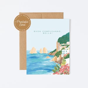 May include: A greeting card featuring a painted coastal scene with blue water, rocky cliffs, and white buildings. The text reads "BUON COMPLEANNO, BELLA!" in dark blue. A circular brown badge in the top left corner says "PRINTABLE CARD". A brown envelope is behind the card.