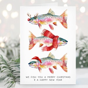 May include: A Christmas card featuring watercolor illustrations of rainbow trout decorated with festive accessories. One fish wears reindeer antlers and a red nose, another a scarf, and the last a Santa hat. The text reads "We fish you a Merry Chiristmas & a Happy New Year."