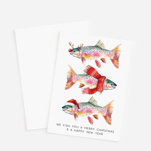 May include: A set of two white greeting cards. The front card features three watercolor fish, each adorned with festive holiday accessories: antlers, a scarf, and a Santa hat. The text reads, "We fish you a Merry Chirstmas & a Happy New Year."