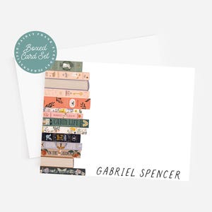 May include: A boxed card set featuring a stack of illustrated books. The book spines have various colors and designs. The card set includes a white card with the name "GABRIEL SPENCER" in black script and a round teal sticker that says "BOXED CARD SET".