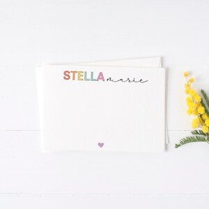 May include: A white card with the name "STELLA marie" written in colorful letters and a small purple heart at the bottom. A sprig of yellow mimosa flowers is on the right side of the card.