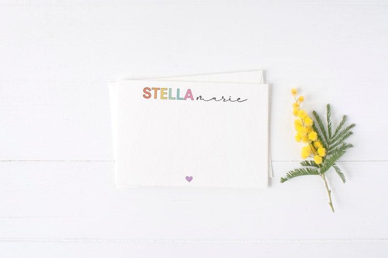 May include: A white card with the name "STELLA marie" written in colorful letters and a small purple heart at the bottom. A sprig of yellow mimosa flowers is on the right side of the card.