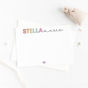 May include: A white card with the name "STELLA marie" written in rainbow letters. A small purple heart is at the bottom of the card.
