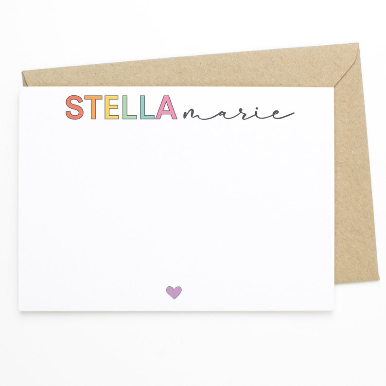 May include: A white card with the name "STELLA marie" written in rainbow colors. A small purple heart is at the bottom of the card.