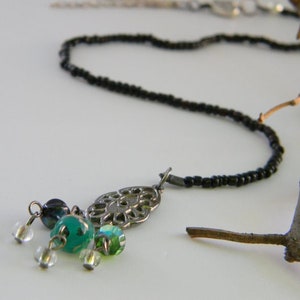 May include: A delicate necklace featuring a black beaded chain and a silver-toned pendant with a filigree design. The pendant is adorned with teal and clear glass beads, creating a subtle contrast. The necklace is displayed on a white surface.