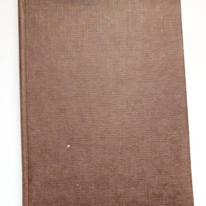 Puede incluir: Un libro cerrado, rectangular, con una cubierta marr&oacute;n texturizada. La cubierta presenta manchas y una ligera decoloraci&oacute;n. El lomo es visible a la izquierda. El libro est&aacute; inclinado sobre un fondo blanco. La cubierta tiene una textura tejida.