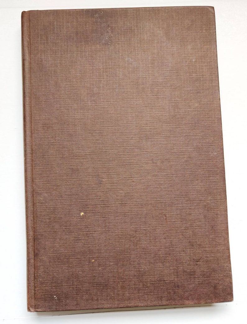 Puede incluir: Un libro cerrado, rectangular, con una cubierta marr&oacute;n texturizada. La cubierta presenta manchas y una ligera decoloraci&oacute;n. El lomo es visible a la izquierda. El libro est&aacute; inclinado sobre un fondo blanco. La cubierta tiene una textura tejida.