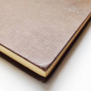 Puede incluir: Primer plano de un libro de tapa dura con una cubierta texturizada de color marr&oacute;n claro. Los bordes de las p&aacute;ginas son dorados y la encuadernaci&oacute;n es de color marr&oacute;n oscuro. El libro est&aacute; sobre un fondo blanco.