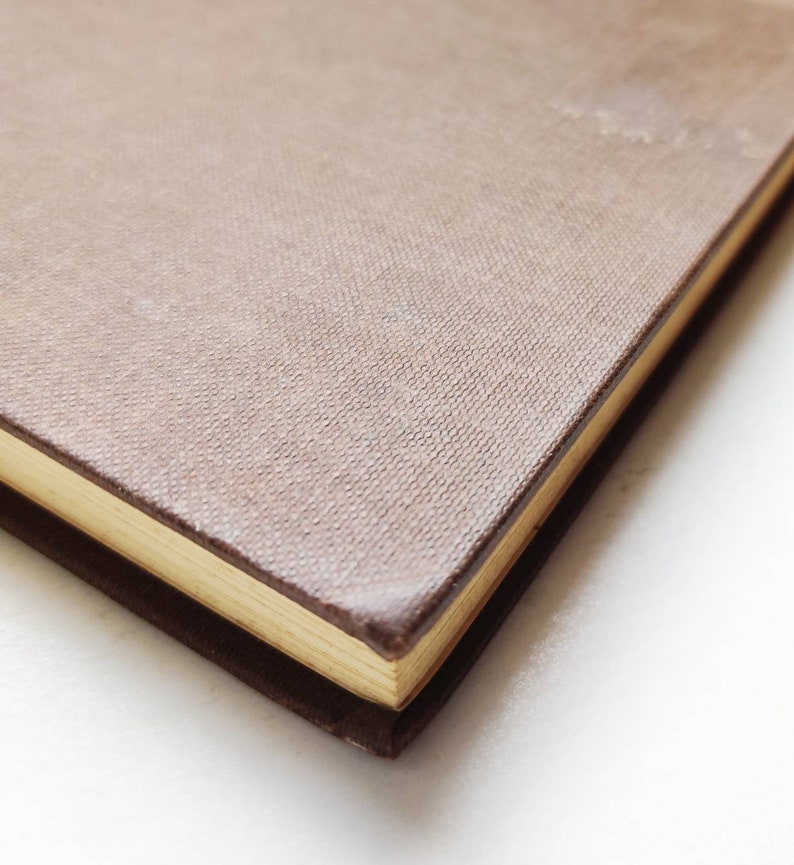 Puede incluir: Primer plano de un libro de tapa dura con una cubierta texturizada de color marr&oacute;n claro. Los bordes de las p&aacute;ginas son dorados y la encuadernaci&oacute;n es de color marr&oacute;n oscuro. El libro est&aacute; sobre un fondo blanco.