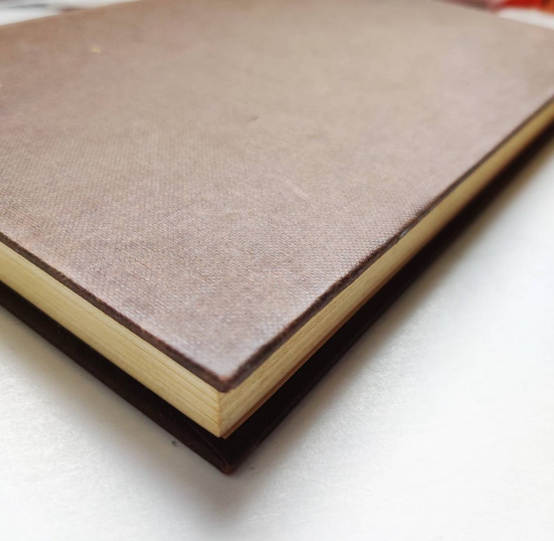 Puede incluir: Primer plano de un diario encuadernado en cuero marr&oacute;n con una cubierta texturizada. Las p&aacute;ginas tienen un tono dorado y los bordes son visibles. El diario descansa sobre una superficie marr&oacute;n m&aacute;s oscura, creando un efecto de capas.