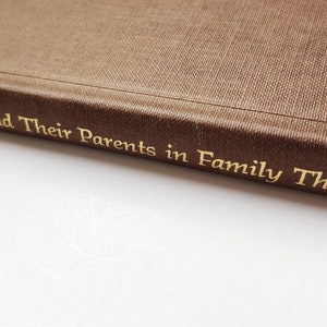 Puede incluir: Primer plano del lomo de un libro marr&oacute;n con letras doradas. El t&iacute;tulo es "Adults and Their Parents in Family Therapy". El libro tiene una cubierta texturizada, probablemente un recurso para terapia familiar.