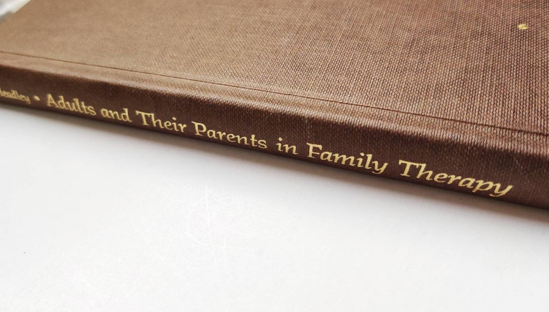 Puede incluir: Primer plano del lomo de un libro marr&oacute;n con letras doradas. El t&iacute;tulo es "Adults and Their Parents in Family Therapy". El libro tiene una cubierta texturizada, probablemente un recurso para terapia familiar.