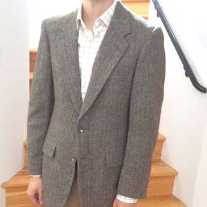 May include: A brown tweed blazer with a herringbone pattern. The blazer has a single button closure and two flap pockets. The blazer is worn over a white button-down shirt with a plaid pattern.