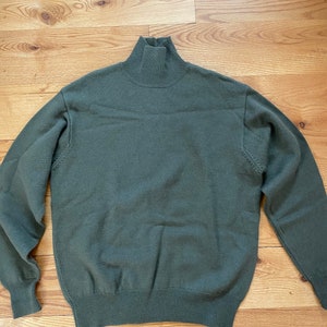 May include: An olive green turtleneck jumper. The jumper has a ribbed collar, cuffs, and hem. The long-sleeved jumper is made of a soft, knitted material. The jumper is laying on a wooden surface.