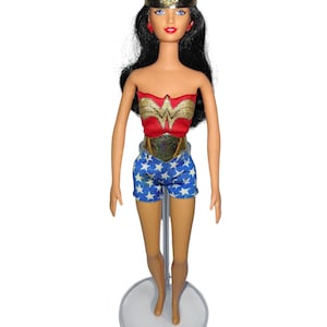 Op de afbeelding: Wonder Woman-pop met een rood en goud lijfje, blauwe shorts met witte sterren en een gouden hoofdband. De pop heeft zwart haar, rode oorbellen en staat op een transparante standaard.