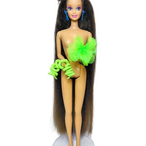 Puede incluir: Una muñeca con cabello largo castaño, pendientes azules y un accesorio de tul verde brillante en el pecho. La muñeca lleva una pulsera de plástico en espiral de color verde lima en un brazo y se sostiene sobre una base circular transparente.