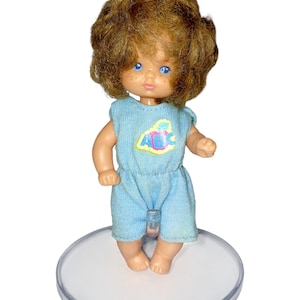 May include: A vintage doll with brown curly hair and blue eyes. The doll is dressed in a light blue romper with a colourful graphic on the chest. It stands on a clear, round display stand. The doll has a vintage aesthetic.