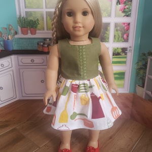 May include: A doll wearing a green sleeveless dress with a white skirt featuring a colorful kitchen utensil pattern. The doll has blonde hair and is wearing red shoes.