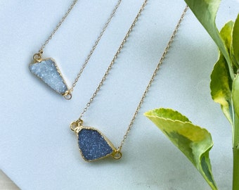 May include: Two gold necklaces with different shaped, irregular, gemstone pendants. The pendants are a light blue and a dark blue. The necklaces are displayed on a white surface with green leaves.