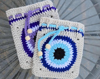 Op de afbeelding: Twee gehaakte trekkoordtassen met boze oogontwerpen. De tassen zijn beige met blauwe, witte en zwarte boze ogen. De tassen hebben paarse en blauwe trekkoorden met houten kralen.