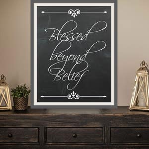 May include: A black framed chalkboard sign with white lettering that reads "Blessed beyond Belief". The sign is sitting on a dark wood dresser with two gold lanterns on either side.