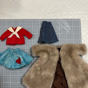 May include: A collection of doll clothes, including a red jacket with a white collar and bow, a blue and black patterned skirt, a light blue skirt with a red heart, and a tan faux fur coat.