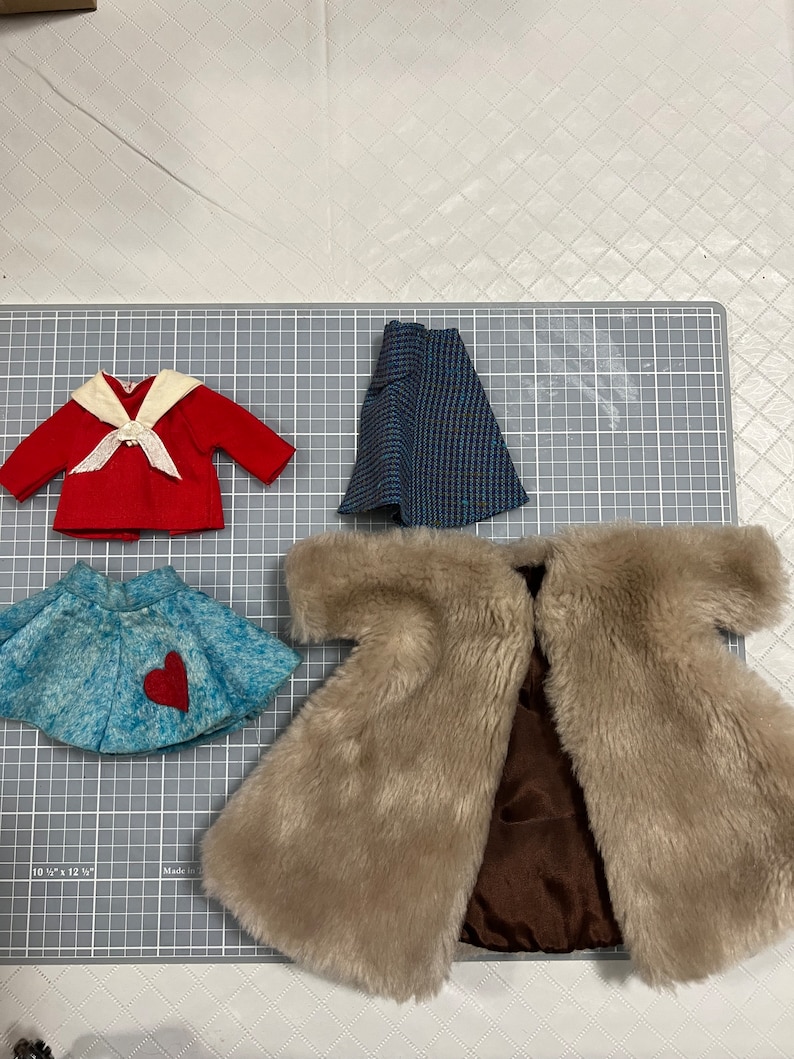 May include: A collection of doll clothes, including a red jacket with a white collar and bow, a blue and black patterned skirt, a light blue skirt with a red heart, and a tan faux fur coat.