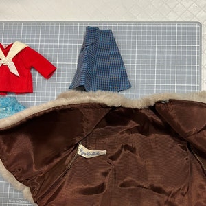 May include: A collection of vintage doll clothes, including a red sailor top with a white collar and bow, a blue and black plaid skirt, and a brown faux fur coat with a satin lining. The coat has a tag that reads "Ginny Doll".