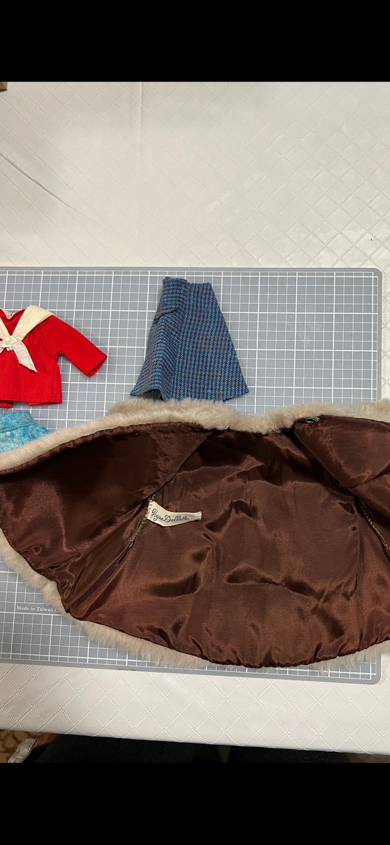 May include: A collection of vintage doll clothes, including a red sailor top with a white collar and bow, a blue and black plaid skirt, and a brown faux fur coat with a satin lining. The coat has a tag that reads "Ginny Doll".