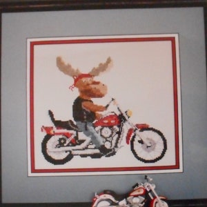 Pode incluir: Um padrão de ponto cruz para um alce a andar de moto. O padrão é intitulado "Biker Moose" e é da empresa Stitchworks. O número de telefone e o endereço da empresa estão incluídos no padrão.