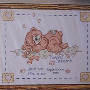 May include: A cross-stitch pattern featuring a brown teddy bear with a smaller teddy bear in its arms. The pattern includes the text "Sweet Dreams" and a personalised message for a baby girl named Jackie Ann, born on 10 September 2012, weighing 6 lbs 10 oz. The pattern is framed in a brown wooden frame.