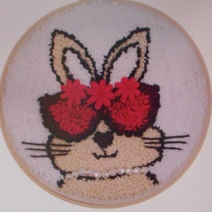 以下が含まれることがあります： パンチニードルキット。直径15.2cmの丸い刺繍枠に、赤いサングラスと真珠のネックレスをした漫画のウサギのデザインが施されています。キットには、プロジェクトを完成させるために必要なすべての材料が含まれています。パッケージには「Loops & Threads」と印刷されています。