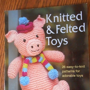 Puede incluir: Un libro titulado "Knitted & Felted Toys" con un cerdo de punto rosa que lleva un gorro azul y rojo y una bufanda a rayas amarilla, rosa y azul. La portada del libro presenta el texto "26 patrones fáciles de tejer para juguetes adorables".