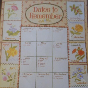 Op de afbeelding: Een vintage-stijl kalender met geborduurde bloemen die elke maand voorstellen. De kalender toont de namen van de maanden en de data om te onthouden, met een bloemmotief voor elke maand.