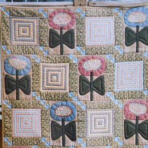 May include: A quilt with a patchwork design featuring a repeating pattern of stylized flowers with bows. The quilt is made with a variety of fabrics in shades of green, brown, pink, blue, and white. The quilt is hanging on a wooden fence with a bush of yellow flowers in the foreground.