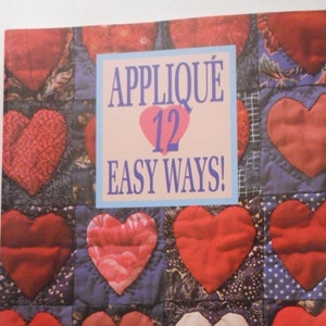 May include: A book cover with a patchwork quilt design featuring red hearts. The title is "Appliqué 12 Easy Ways!" with a red heart around the number 12. The book is by Elly Sienkiewicz and describes charming quilts, giftable projects, and timeless techniques.